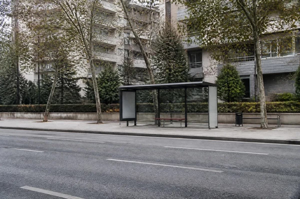 Cadde kenarında camlı otobüs durağı barınağı; arkada çıplak ağaçlar ve apartmanlar